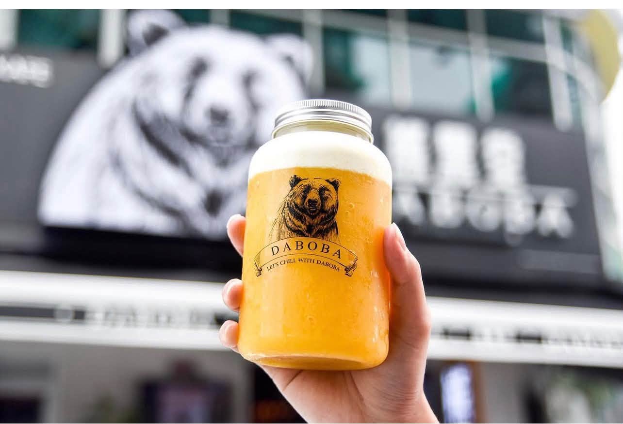 A customer holding a fresh mango Daboba drink in front of the bear mural outside the store